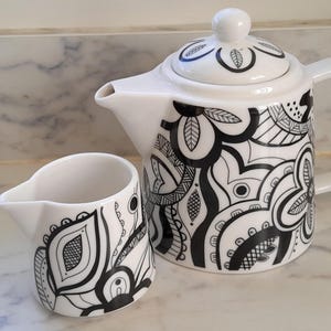 May include: A white ceramic teapot and creamer set, decorated with black abstract floral patterns. The teapot has a lid and handle, and the creamer has a spout and handle. The set is on a marble countertop.