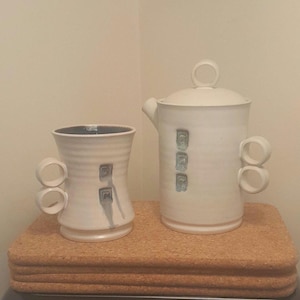 Może przedstawiać: Biały ceramiczny czajnik i kubek z niebieskimi i szarymi akcentami. Czajnik ma pokrywkę z uchwytem w kształcie pierścienia i dwa boczne uchwyty. Kubek ma dwa uchwyty i zaokrąglony kształt.