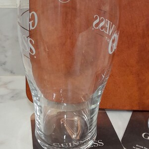 May include: A clear Guinness pint glass with the Guinness logo and text. The glass is on a coaster with the Guinness logo and text. The glass has a capacity of 20 fluid ounces.