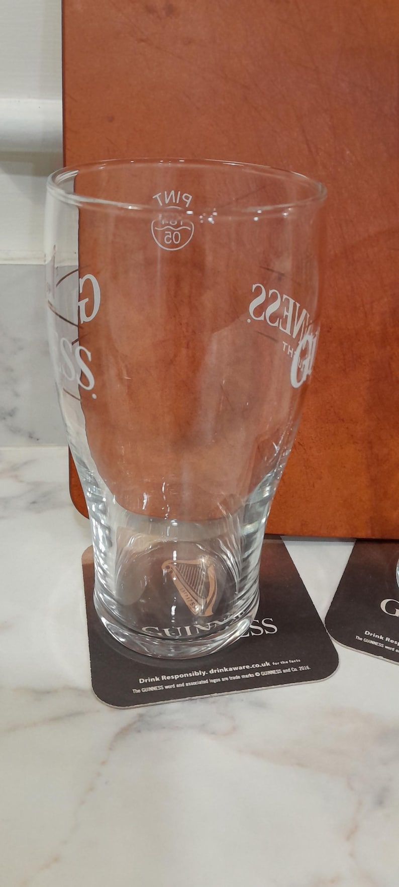 May include: A clear Guinness pint glass with the Guinness logo and text. The glass is on a coaster with the Guinness logo and text. The glass has a capacity of 20 fluid ounces.