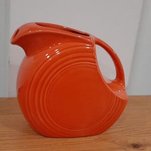 Może przedstawiać: Jasnopomarańczowy dzbanek ceramiczny o zaokrąglonym korpusie z koncentrycznymi, wytłoczonymi pierścieniami, zakrzywionym uchwytem i wyraźnym dzióbkiem. Dzbanek ma gładkie, błyszczące wykończenie i charakterystyczny kształt przypominający dysk.