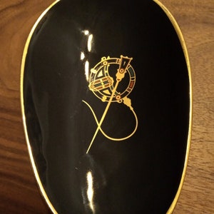 May include: A black oval dish with a gold rim and a gold design of a stylized sword and circle with red, green, and gold accents.