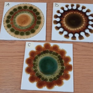 May include: Three square white ceramic tiles labeled A, B, and C. Tile A features a circular pattern of green and tan rings. Tile B displays a dark brown sunburst design with white accents. Tile C shows a central green circle with orange petals.