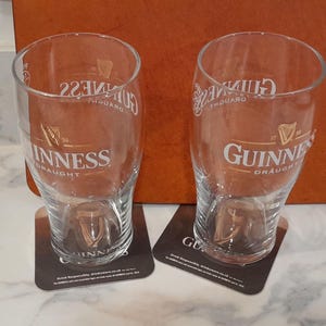 May include: Two clear Guinness pint glasses with the Guinness logo and text, resting on square coasters. The glasses are empty and sit on a white marble surface. The background includes a brown surface.