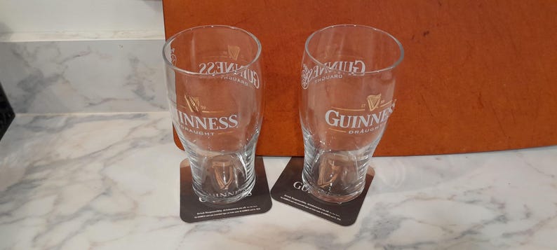May include: Two clear Guinness pint glasses with the Guinness logo and text, resting on square coasters. The glasses are empty and sit on a white marble surface. The background includes a brown surface.