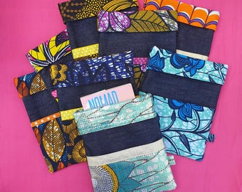Puede incluir: Una colección de bolsas de mezclilla con coloridos detalles de tela estampada. Las bolsas presentan varios diseños, incluyendo patrones florales, geométricos y abstractos en tonos de azul, amarillo, naranja y morado. Una bolsa contiene una tarjeta rosa con la palabra "NOMAD" impresa.