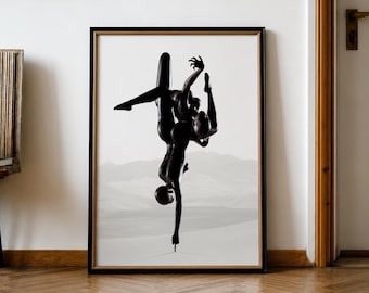 May include: Black and white framed art print featuring a stylized depiction of two figures in a dynamic pose against a desert landscape. The artwork is displayed on a white wall, complemented by a wooden frame and a parquet floor.