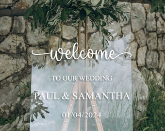 May include: A clear acrylic wedding sign with the text "Welcome to our wedding Paul & Samantha 01/04/2024" in white lettering. The sign is decorated with a floral arrangement of white roses and green leaves.