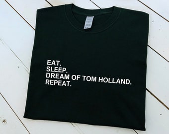 May include: A dark green t-shirt with white text that reads "EAT. SLEEP. DREAM OF TOM HOLLAND. REPEAT."