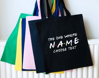 May include: Black tote bag with white text that reads "The One Where NAME CHOOSE TEXT". The text is in a font similar to the Friends logo. The bag is shown with other tote bags in yellow, pink, blue, and green.