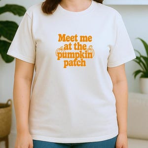 May include: A cream-coloured t-shirt featuring the orange text "Meet me at the pumpkin patch". The shirt is worn with blue jeans. The image evokes a sense of autumn and fall.
