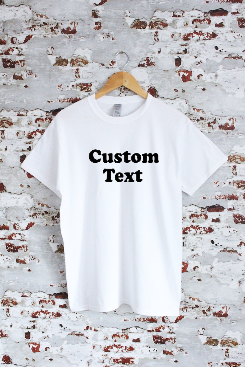 May include: A white t-shirt with the words "Custom Text" printed in bold black letters. The shirt is hanging on a wooden hanger against a textured brick wall background.