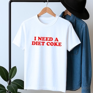 May include: White t-shirt with the red text "I NEED A DIET COKE." The shirt is hanging on a black clothing rack with a denim shirt and black hat in the background. A plant is visible on the left.