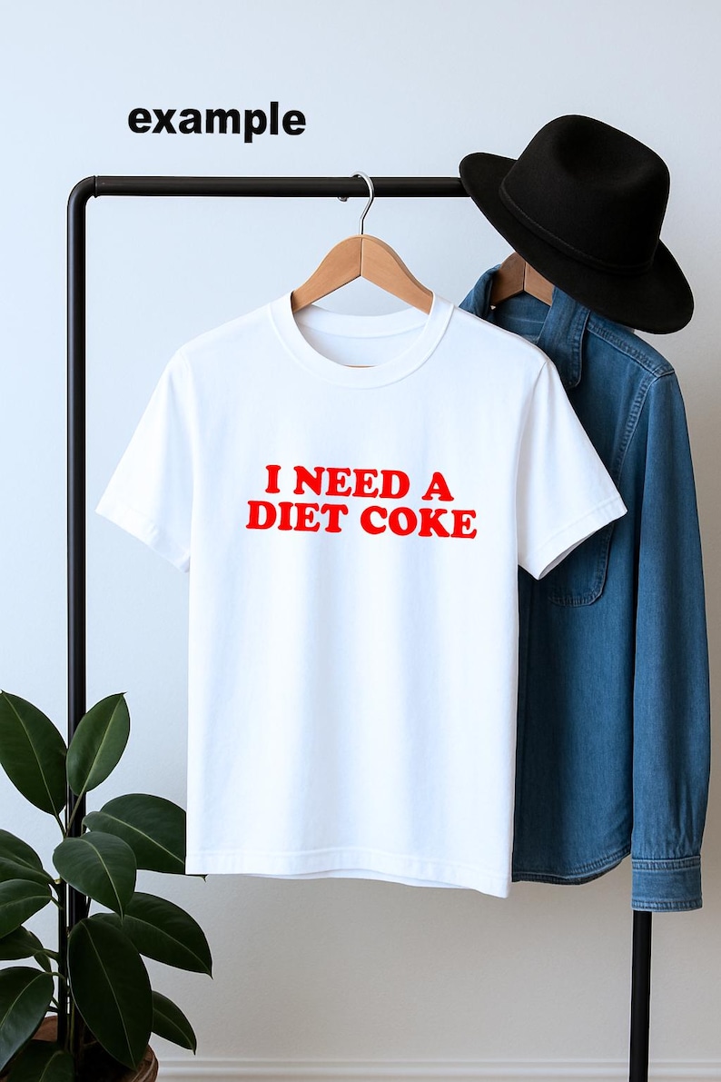 May include: White t-shirt with the red text "I NEED A DIET COKE." The shirt is hanging on a black clothing rack with a denim shirt and black hat in the background. A plant is visible on the left.