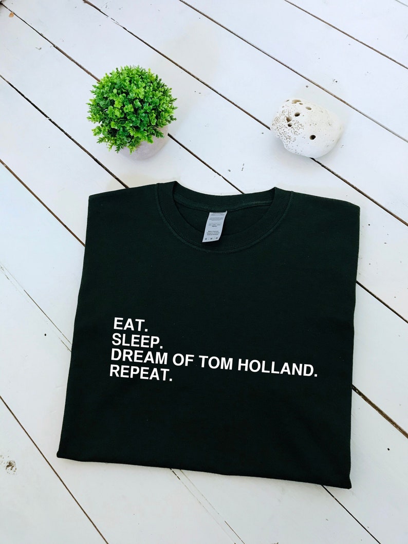 May include: A dark green t-shirt with white text that reads "EAT. SLEEP. DREAM OF TOM HOLLAND. REPEAT."