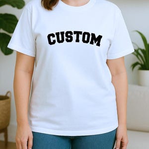 May include: A white t-shirt with the word "CUSTOM" printed in bold, black letters across the chest. The model is wearing blue jeans. The shirt has short sleeves and a crew neck.