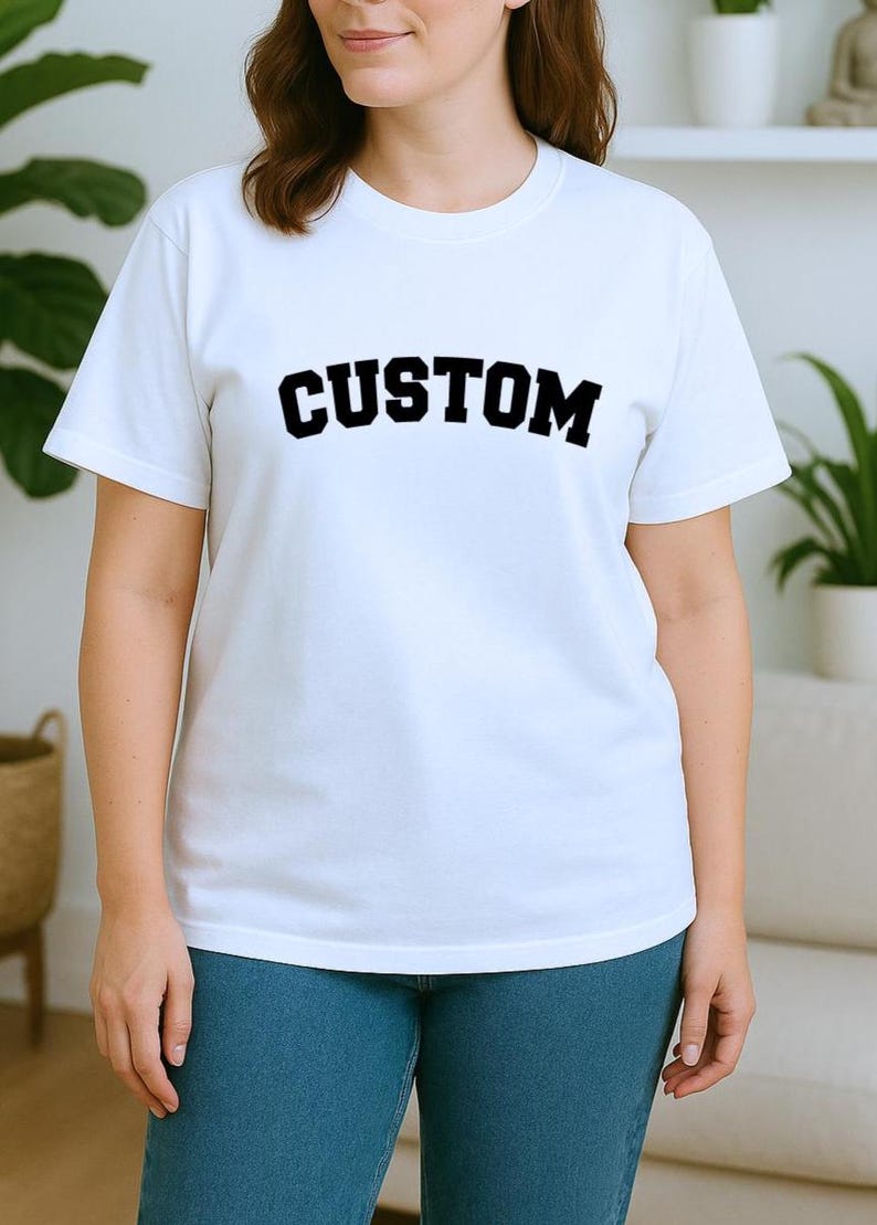 May include: A white t-shirt with the word "CUSTOM" printed in bold, black letters across the chest. The model is wearing blue jeans. The shirt has short sleeves and a crew neck.