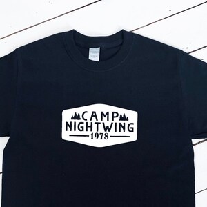 May include: Black t-shirt with a white graphic of a camp logo. The logo says "Camp Nightwing - 1978".