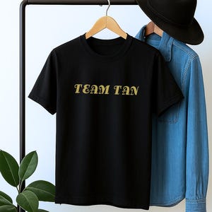May include: Black crew neck t-shirt with the text "TEAM TAN" in gold lettering. A denim shirt and black hat hang on a clothing rack behind the t-shirt. The word "example" is above the t-shirt.