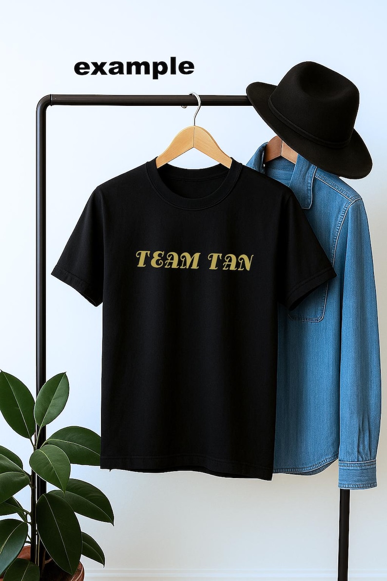 May include: Black crew neck t-shirt with the text "TEAM TAN" in gold lettering. A denim shirt and black hat hang on a clothing rack behind the t-shirt. The word "example" is above the t-shirt.