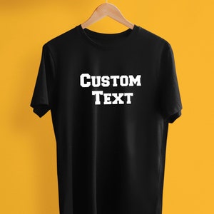 May include: Black t-shirt with the words "CUSTOM TEXT" in bold, white letters. The shirt is hanging on a wooden hanger against a yellow background. The shirt is a classic crew neck style.