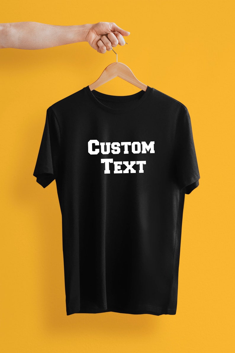 May include: Black t-shirt with the words "CUSTOM TEXT" in bold, white letters. The shirt is hanging on a wooden hanger against a yellow background. The shirt is a classic crew neck style.