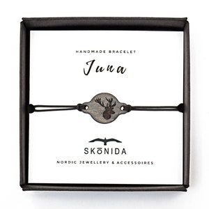 Op de afbeelding: Een zwarte doos met een witte achtergrond. De doos bevat een handgemaakt armband met een houten hertenbedel. Het armband is gemaakt door Sk&ouml;nida, een Scandinavisch bedrijf voor sieraden en accessoires. De doos heeft ook de tekst "Handgemaakt Armband" en "Juna" erop.