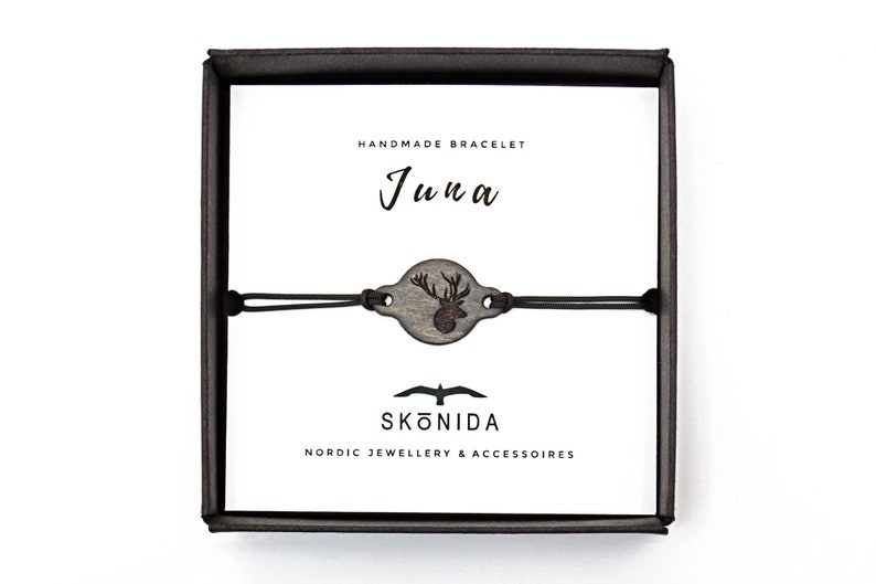 Op de afbeelding: Een zwarte doos met een witte achtergrond. De doos bevat een handgemaakt armband met een houten hertenbedel. Het armband is gemaakt door Sk&ouml;nida, een Scandinavisch bedrijf voor sieraden en accessoires. De doos heeft ook de tekst "Handgemaakt Armband" en "Juna" erop.