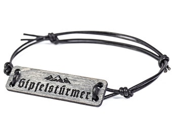 Op de afbeelding: Een zwart leren armband met een houten tag waarop "Gipfelstürmer" staat met een bergketen afbeelding.