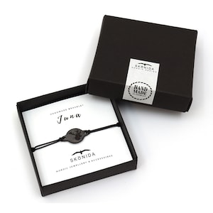 Op de afbeelding: Een zwarte doos met een wit label waarop "HAND MADE IN GERMANY" staat en een zwarte armband met een houten hertenbedel. De armband zit in een zwarte doos met een wit label waarop "Handmade Bracelet" en "Juna" staat.