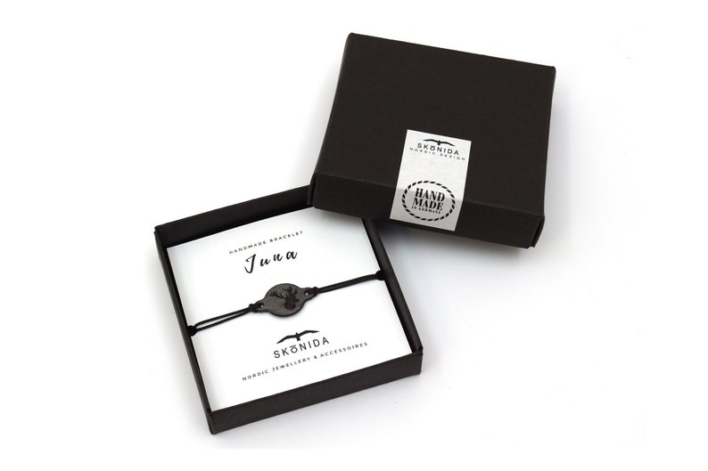 Op de afbeelding: Een zwarte doos met een wit label waarop "HAND MADE IN GERMANY" staat en een zwarte armband met een houten hertenbedel. De armband zit in een zwarte doos met een wit label waarop "Handmade Bracelet" en "Juna" staat.
