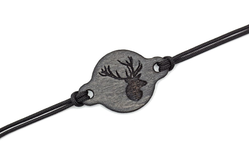 Op de afbeelding: Een zwarte houten armband met een ronde hanger met een gegraveerd hertenkopontwerp. De armband is gemaakt van zwart koord.