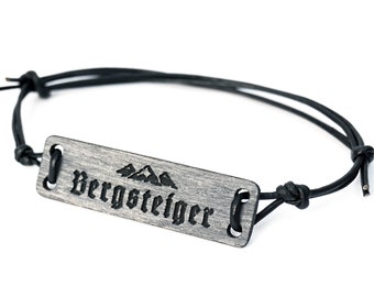 Op de afbeelding: Een zwart leren armband met een rechthoekig zilveren label met de tekst "Bergsteiger" en een afbeelding van een bergketen.