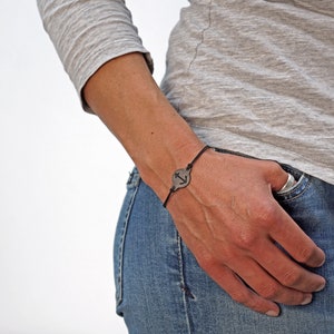 Op de afbeelding: Een armband van zwart koord met een ronde zilveren bedel met een ankerontwerp.