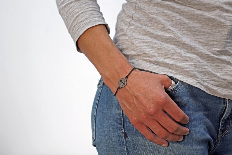 Op de afbeelding: Een armband van zwart koord met een ronde zilveren bedel met een ankerontwerp.