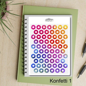 May include: A sheet of colorful circle stickers arranged in rows, transitioning from pink to orange and blue to purple. The sheet is labeled "Konfetti 1" and rests on a white pad atop a green notebook. A plant and pens are in the background.