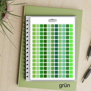 May include: A spiral-bound notebook with a white page featuring a grid of green squares in varying shades. The word "HARZPLOTT" is printed at the top. The word "grün" is printed at the bottom. The notebook rests on a green book.