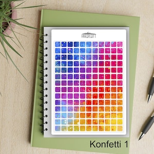 May include: A sheet of colorful square stickers arranged in a gradient from blue to yellow, with the text "Konfetti 1" at the bottom. The stickers are on a white background, atop a spiral-bound notebook and a green book.