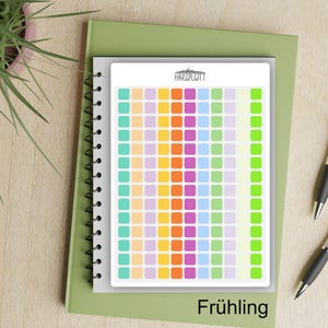 May include: A colorful planner with a grid of small squares in various colors, including yellow, purple, and green. The planner is on a green notebook, with the word "Frühling" at the bottom. Two pens are visible.