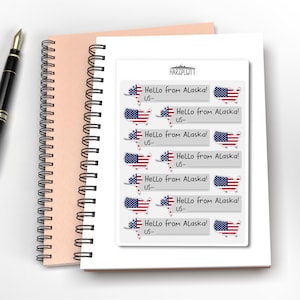 May include: A sheet of stickers with the text "Hello from Alaska!" and a map of Alaska in the colors of the American flag. The stickers are on a white background, with a spiral-bound notebook and a pen in the background.