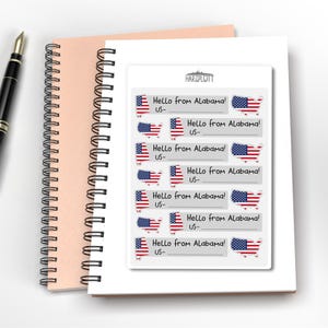 May include: A stack of stickers with the text "Hello from Alabama!" and a US flag design. The stickers are on a white background, with a pink notebook and a pen in the background.