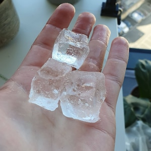 May include: Four clear ice cubes on a hand. The ice cubes are square shaped and have a frosted appearance.
