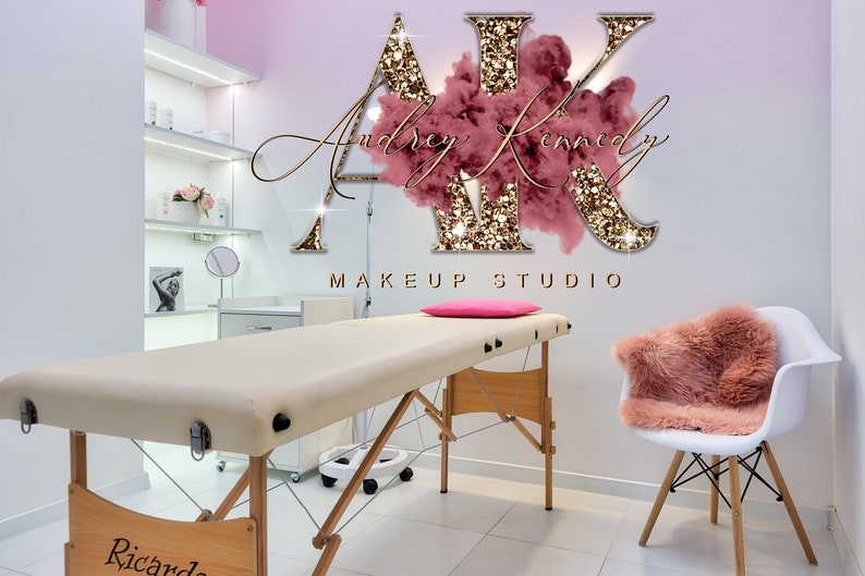 May include: A makeup studio with a white chair and a beige treatment table. The wall has a pink and gold glitter logo that says "AK Makeup Studio" with the text "Audrey Kennedy" in cursive.