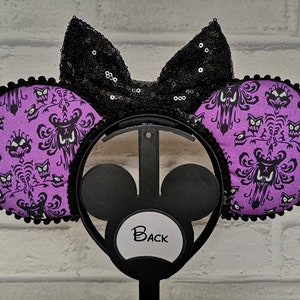 May include: Purple and black Minnie Mouse ears with a black bow and a Haunted Mansion wallpaper pattern. The ears are on a black headband with a white label that says "Back".