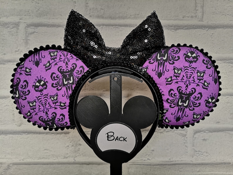 May include: Purple and black Minnie Mouse ears with a black bow and a Haunted Mansion wallpaper pattern. The ears are on a black headband with a white label that says "Back".