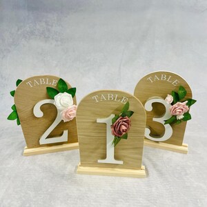 May include: Three wooden table number signs with floral accents. Each sign has a light wood grain finish, a white number, and the word "TABLE" at the top. The numbers are 1, 2, and 3. The signs are decorated with white, pink, and burgundy flowers.
