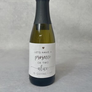 May include: A small glass bottle of prosecco with a black foil-wrapped neck and a white rectangular label. The label features a small heart icon and the text: LETS HAVE A prosecco OR TWO alice IS SAYING I DO in black and grey lettering.