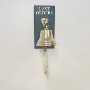 May include: A polished brass bell mounted on a dark rectangular wooden plaque with the text LAST ORDERS printed in white capital letters. The bell features a braided white rope pull attached to the clapper, hanging vertically from the wall mount.