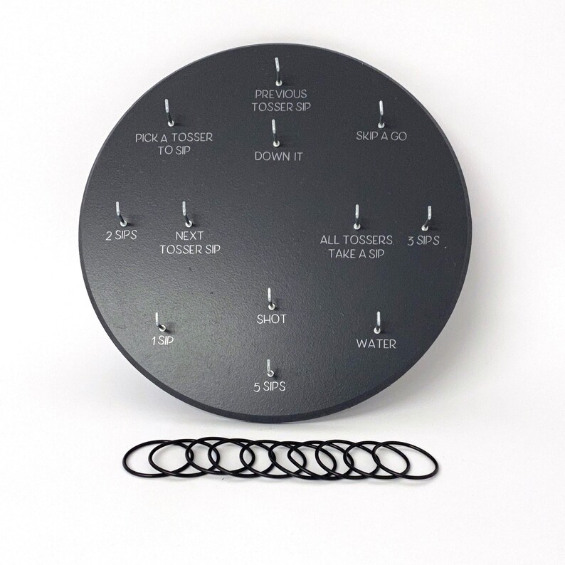 May include: A black circular game board with 10 metal hooks and corresponding text labels. The labels include "Pick a Tosser to Sip", "Previous Tosser Sip", "Down It", "Skip a Go", "2 Sips", "Next Tosser Sip", "All Tossers Take a Sip", "3 Sips", "1 Sip", "Shot", "Water", and "5 Sips".  There are 10 black rubber rings below the board.