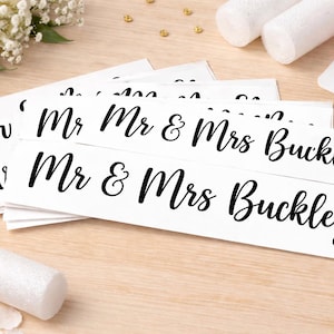 Puede incluir: Pegatinas rectangulares blancas con texto negro en cursiva "Mr & Mrs Buckley". Las pegatinas están apiladas sobre una superficie de madera clara, con decoración de boda.